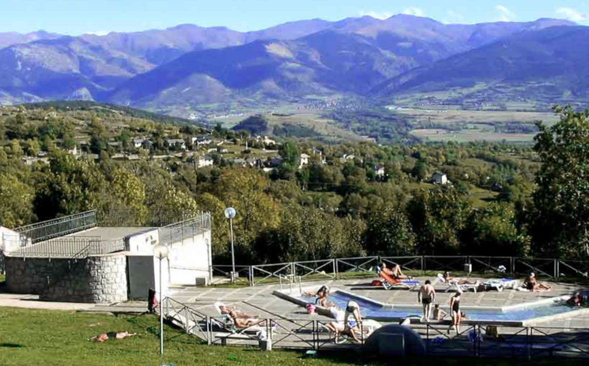 Un espace piscine comprenant plusieurs bassins, entouré d’un bâtiment en pierre, le tout situé dans un cadre montagneux.