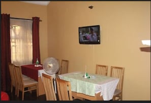 Zona de menjador amb una taula preparada per a quatre, un televisor fixat a la paret i una finestra amb cortines.