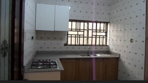 A compact kitchen with a gas stove, wooden cabinets, a window with bars, and patterned wall tiles. - Cristabol Apartments (Lekki)