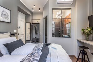 A modern bedroom with a large bed, a walk-in closet, a bathroom with a glass shower, and a view of a brick wall. - Deluxe Ploech (Rijeka)