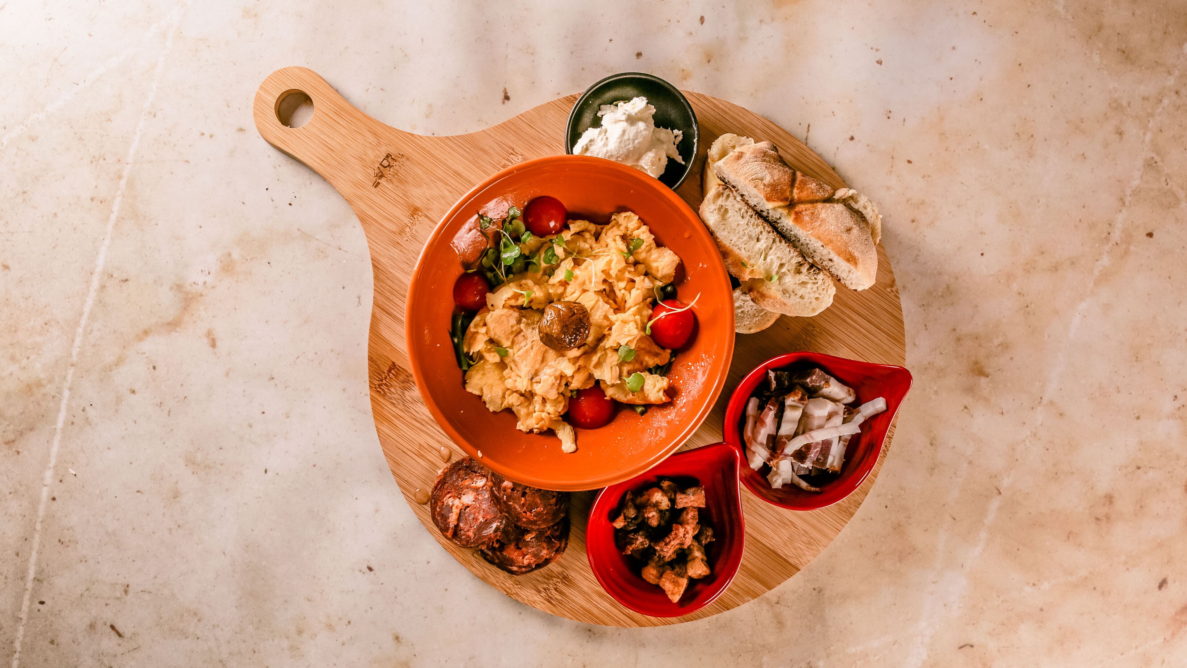 A wooden serving board with scrambled eggs, cherry tomatoes, and greens, accompanied by bread and a bowl of meat.