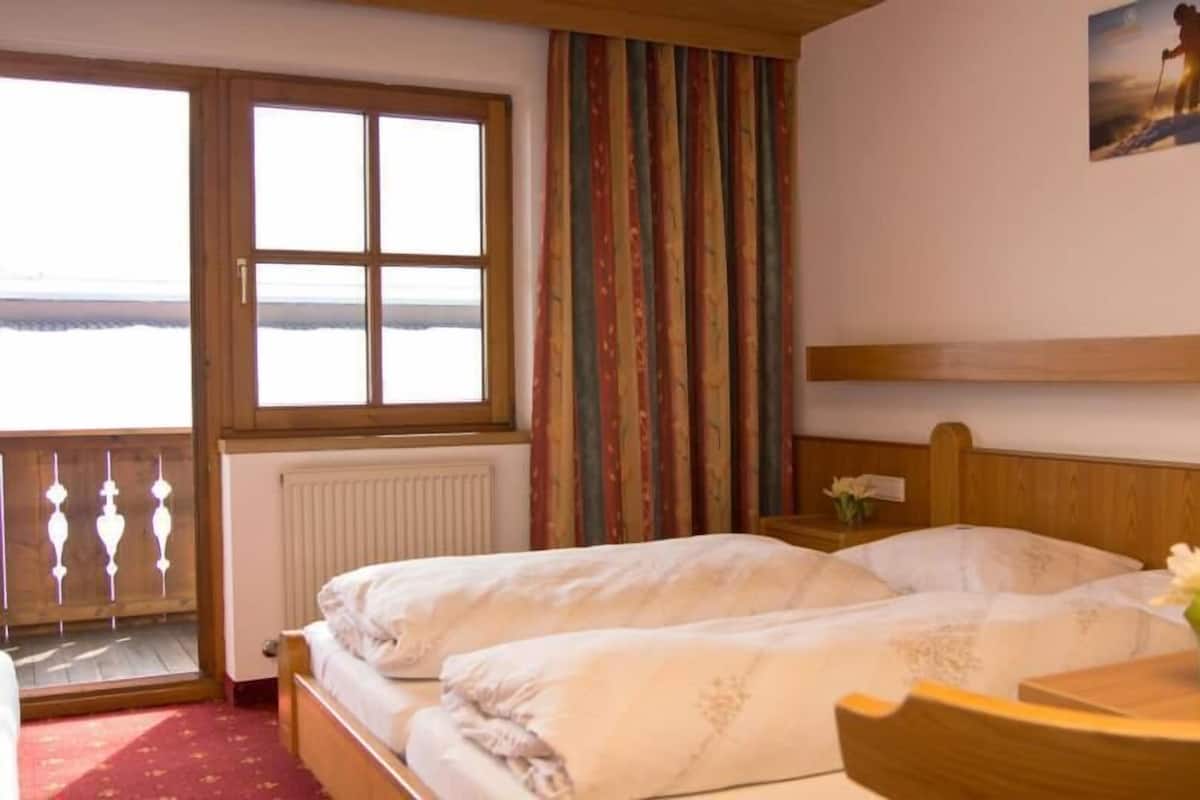 A hotel room with a bed, a window with a view, a wooden headboard, and a small table with a vase of flowers.