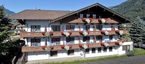 A multi-story hotel with balconies adorned with flower baskets, set against a mountainous backdrop.