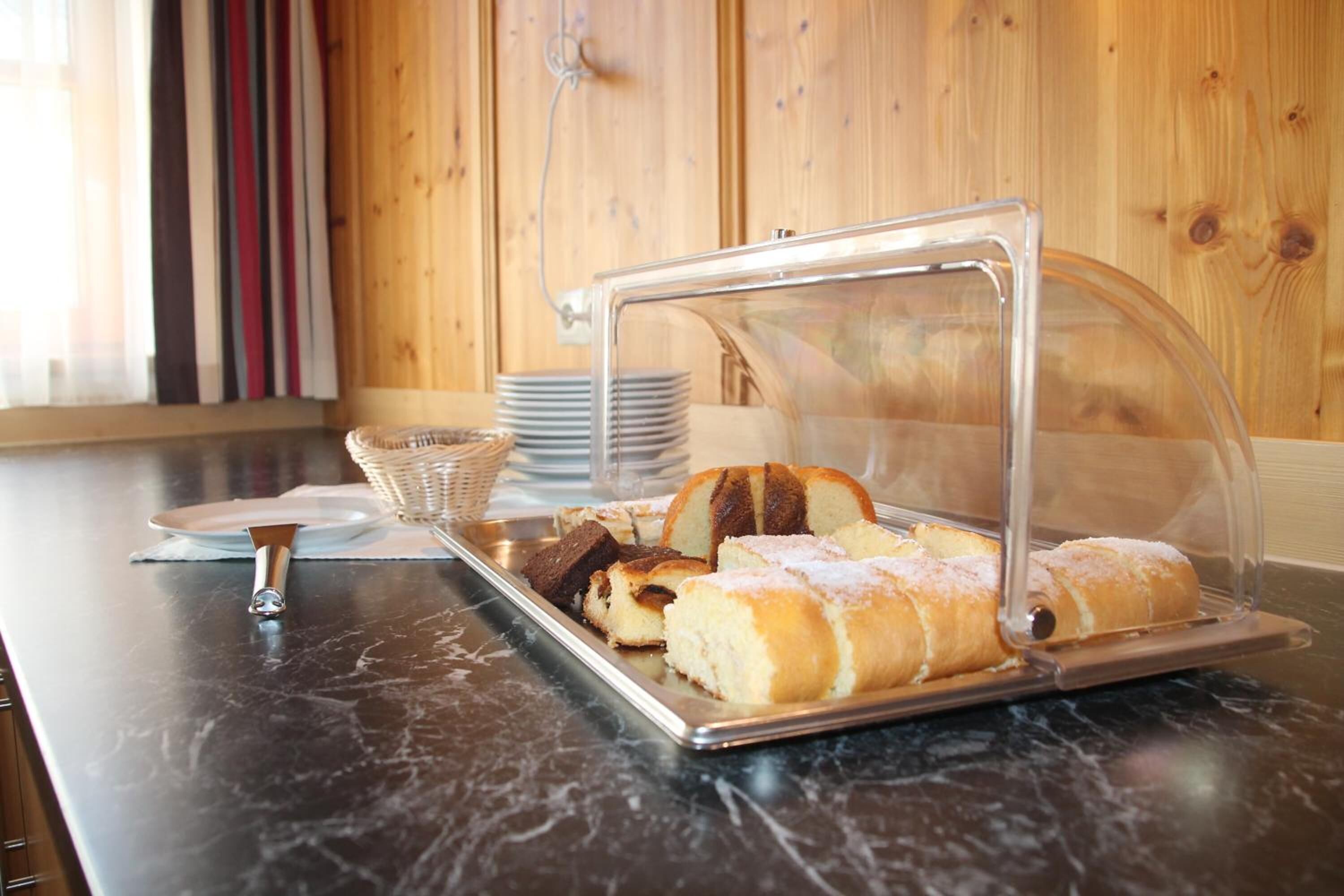 A buffet setup with a clear glass cover, a tray of bread rolls, and a stack of plates on a marble countertop.