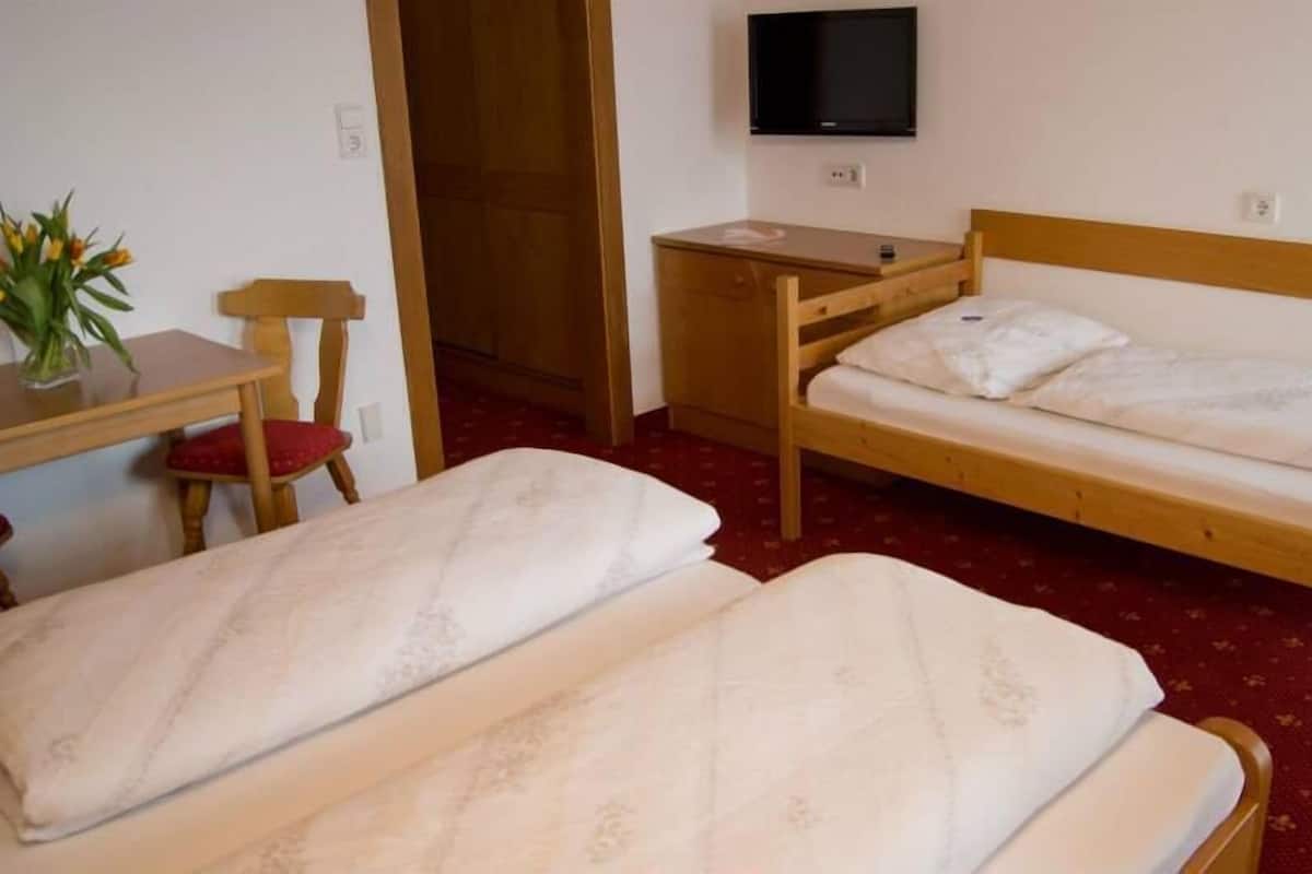 A room with two single beds, a wooden desk with a chair, a TV mounted on the wall, and a small table with a vase of flowers.
