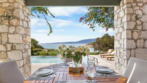 Un espace de restauration en plein air, avec des murs en pierre, une table en bois dressée pour deux personnes et une vue sur la mer.