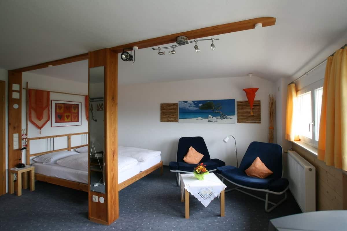Une pièce avec un lit en bois, une petite table sur laquelle est posé un vase, deux chaises et un tableau accroché au mur.
