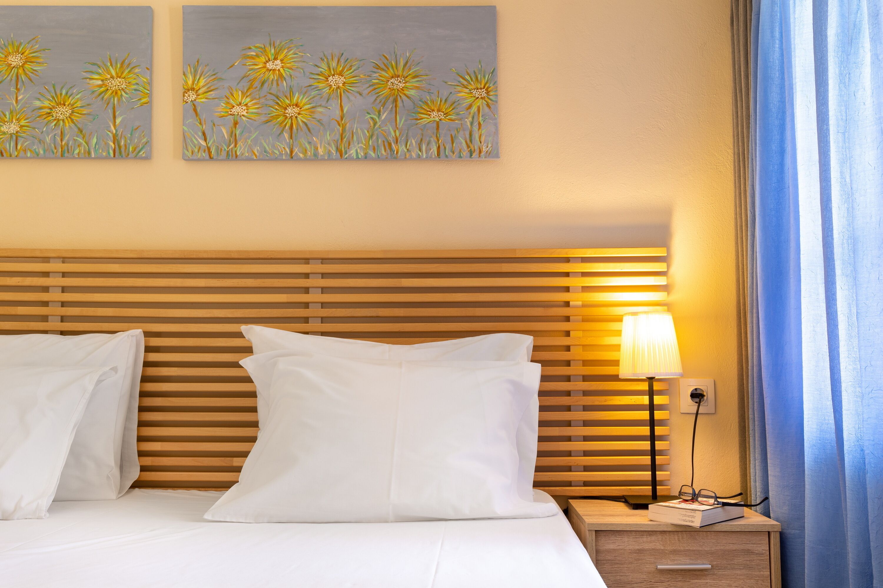 A hotel room with a wooden headboard, white bedding, two white pillows, a bedside table with a lamp, and two paintings of flowers on the wall.