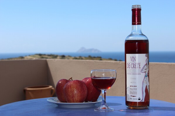 Una botella de vino Vin de Crete, una copa de vino tinto y tres manzanas rojas sobre una mesa con vista al mar y una montaña al fondo.