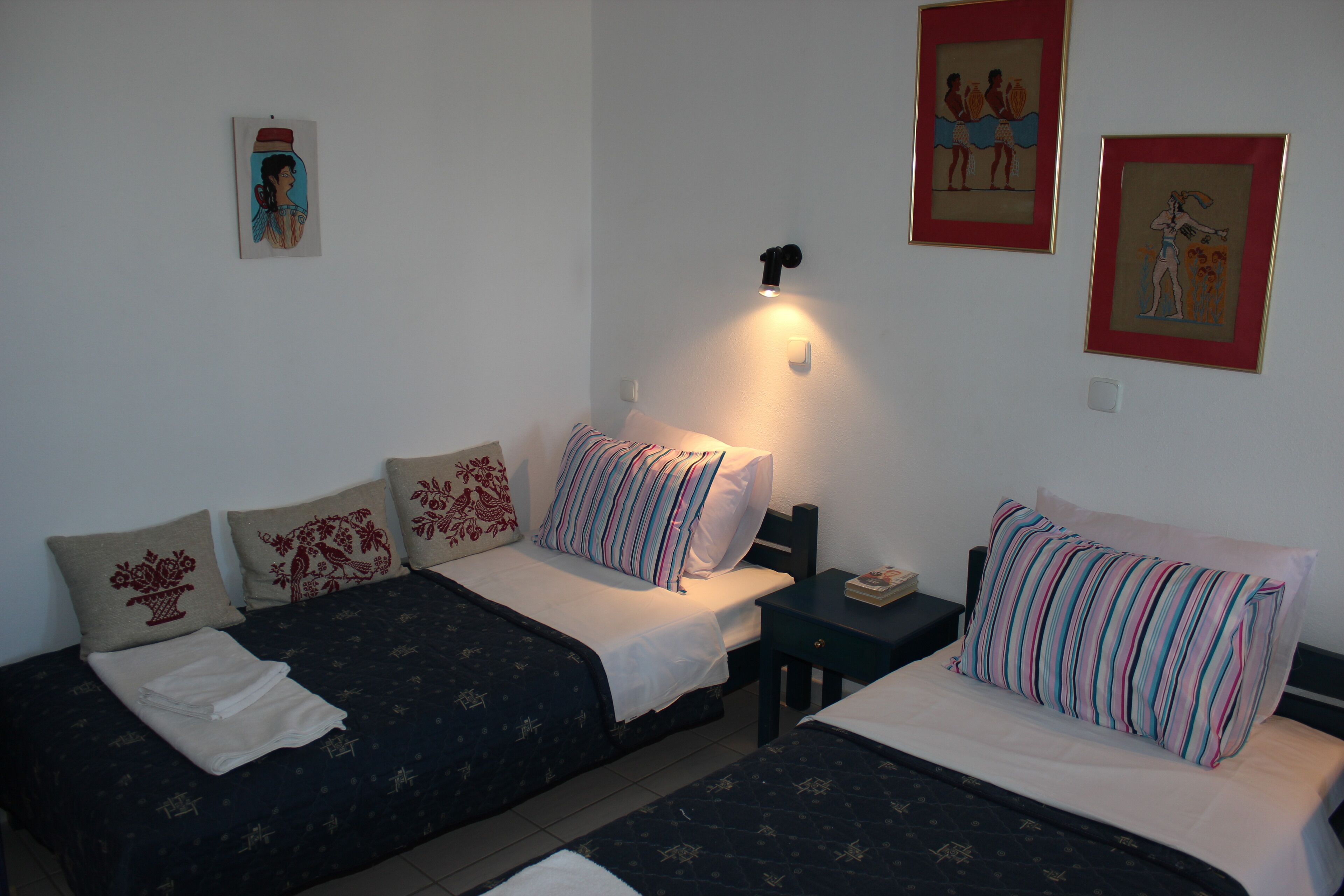 Habitación de hotel con dos camas, cada una con un cojín a rayas y uno decorativo. Hay una mesita de noche con una lámpara entre las camas.
