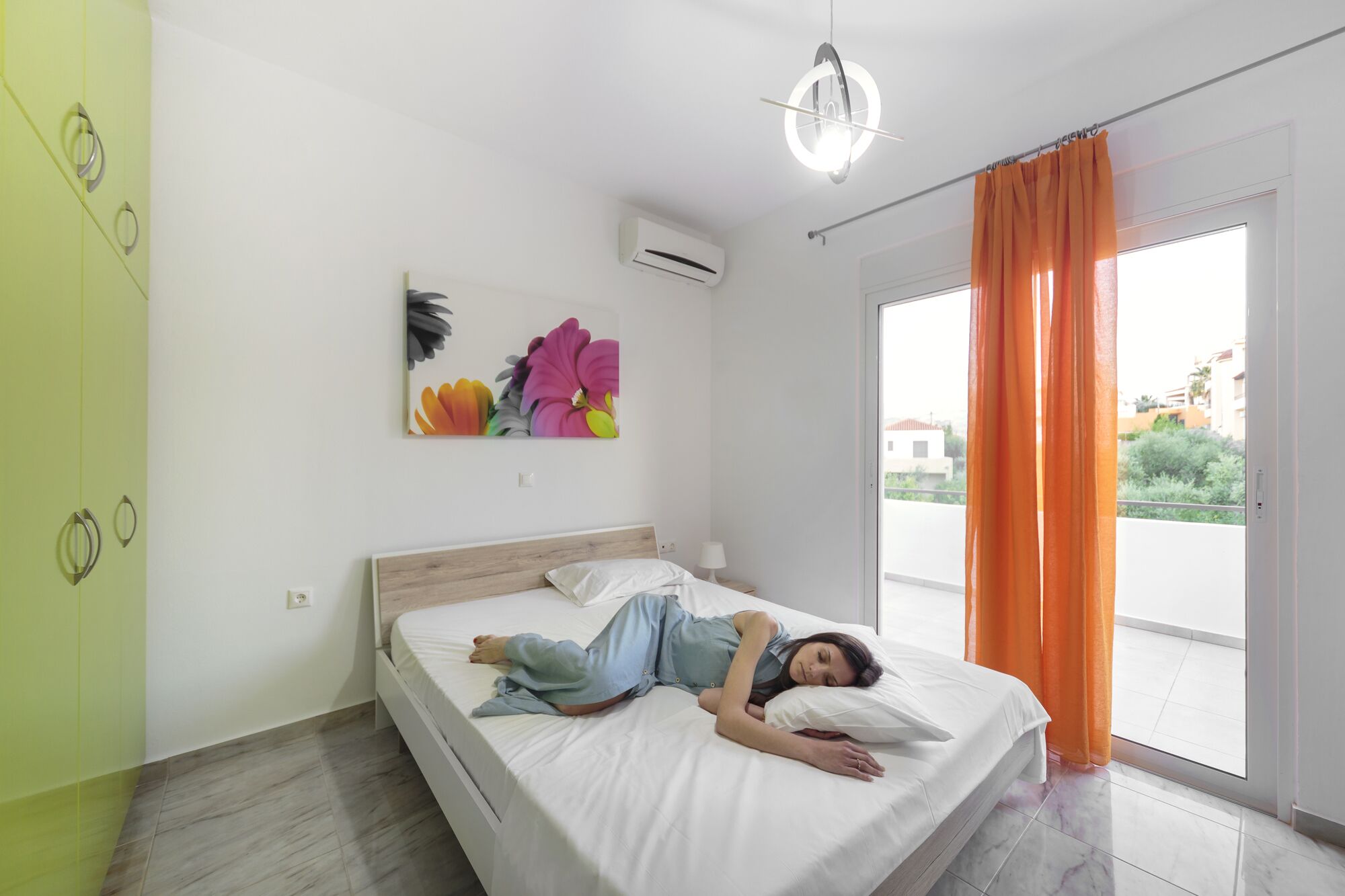 Ein Schlafzimmer mit einem Bett, einer Wand-Klimaanlage, einem Fenster mit orangefarbenen Vorhängen und Blick ins Freie.