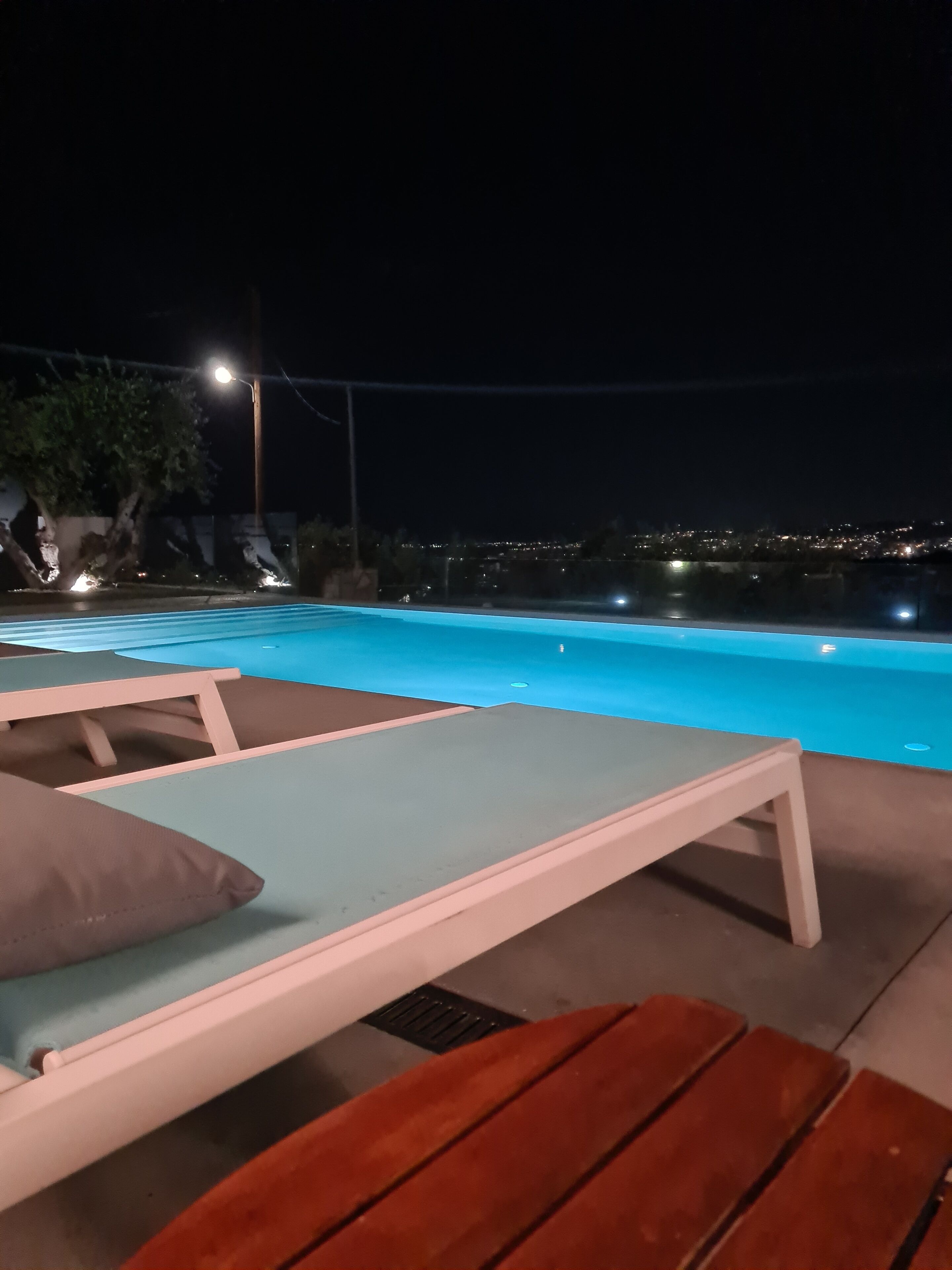 Un espace aménagé au bord de la piscine, avec des chaises longues et une table, entouré d’arbres et offrant une vue sur le paysage urbain illuminé la nuit.