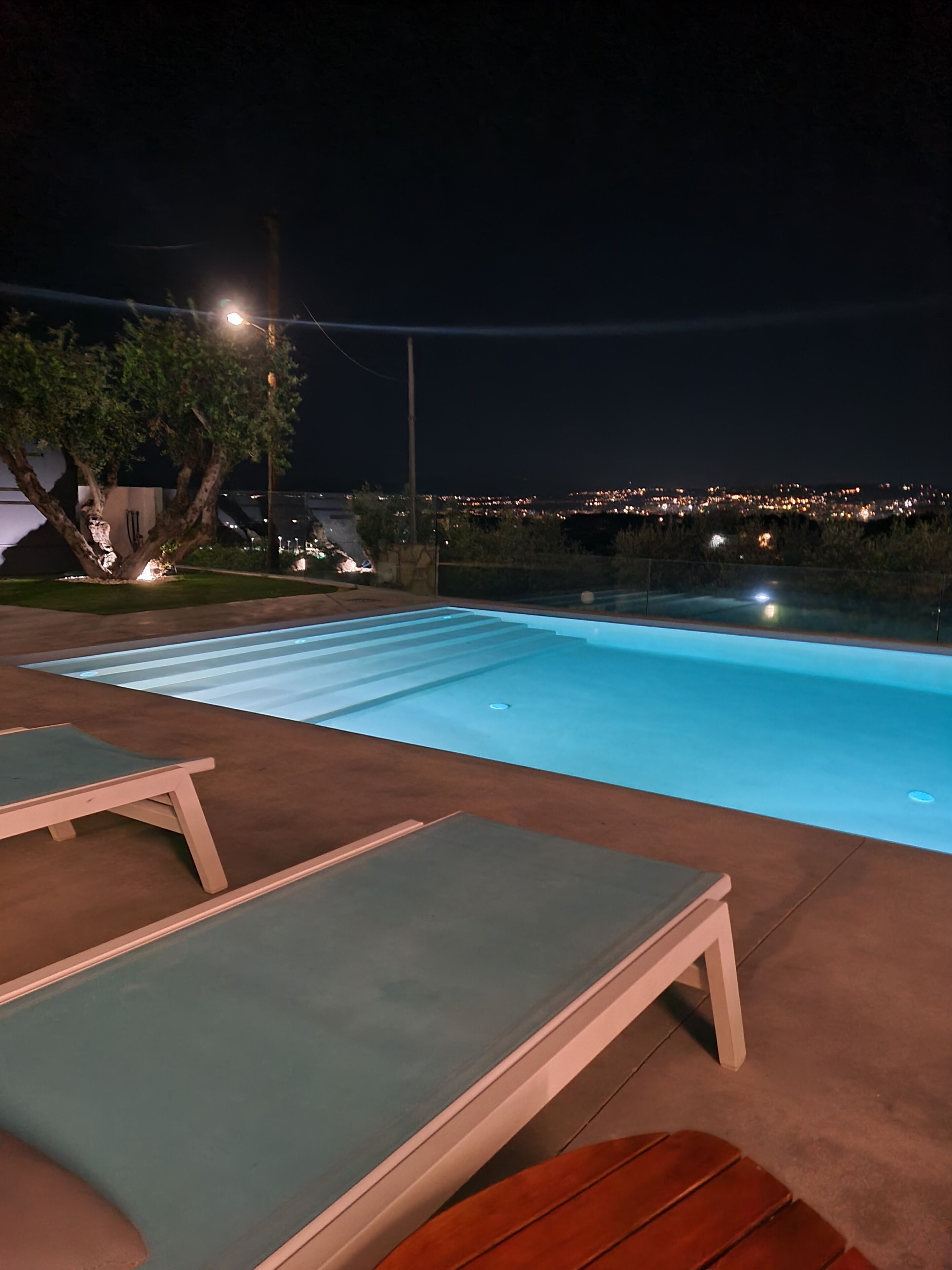 An outdoor swimming pool with lounge chairs, a tree, and a cityscape view at night.