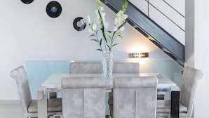 Ein moderner Essbereich mit einem Glastisch, grau gepolsterten Stühlen und einer Vase mit weißen Blumen. An der Wand befinden sich schwarze Schallplatten und im Hintergrund eine Treppe.