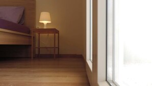 Quarto com piso de madeira, cama com travesseiro roxo e mesa de cabeceira com abajur.