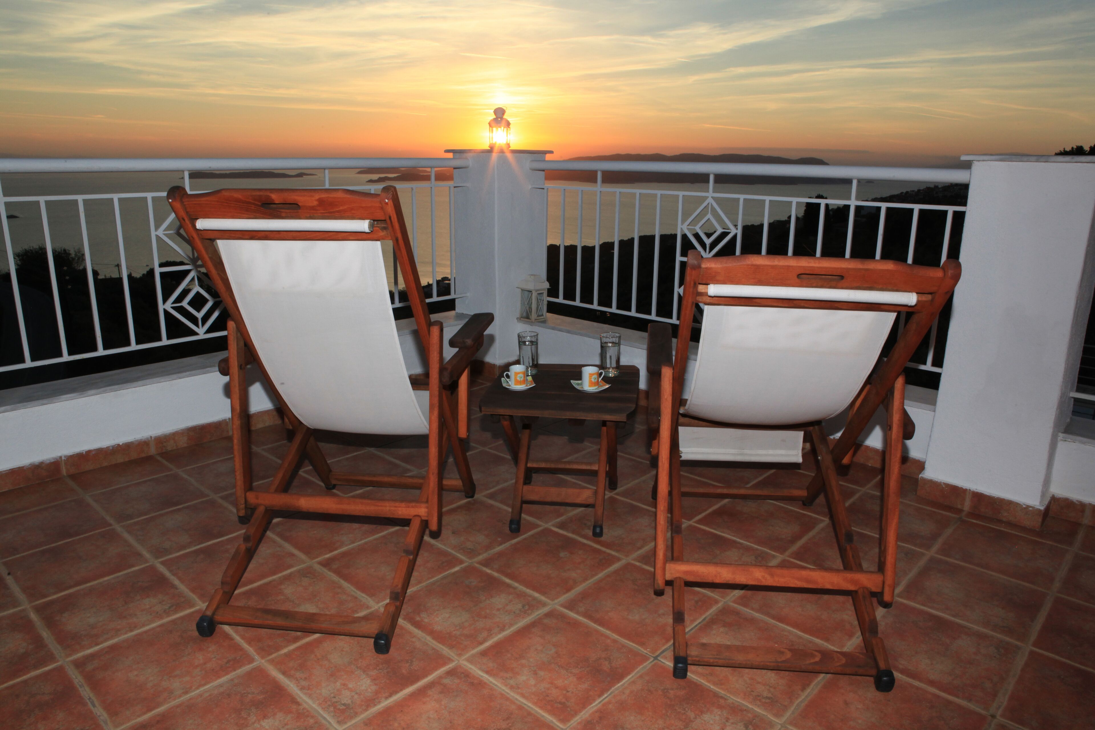 A balcony with two wooden chairs and a small table, overlooking a sunset view.