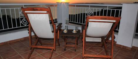 A balcony with two wooden chairs and a small table, overlooking a sunset view.