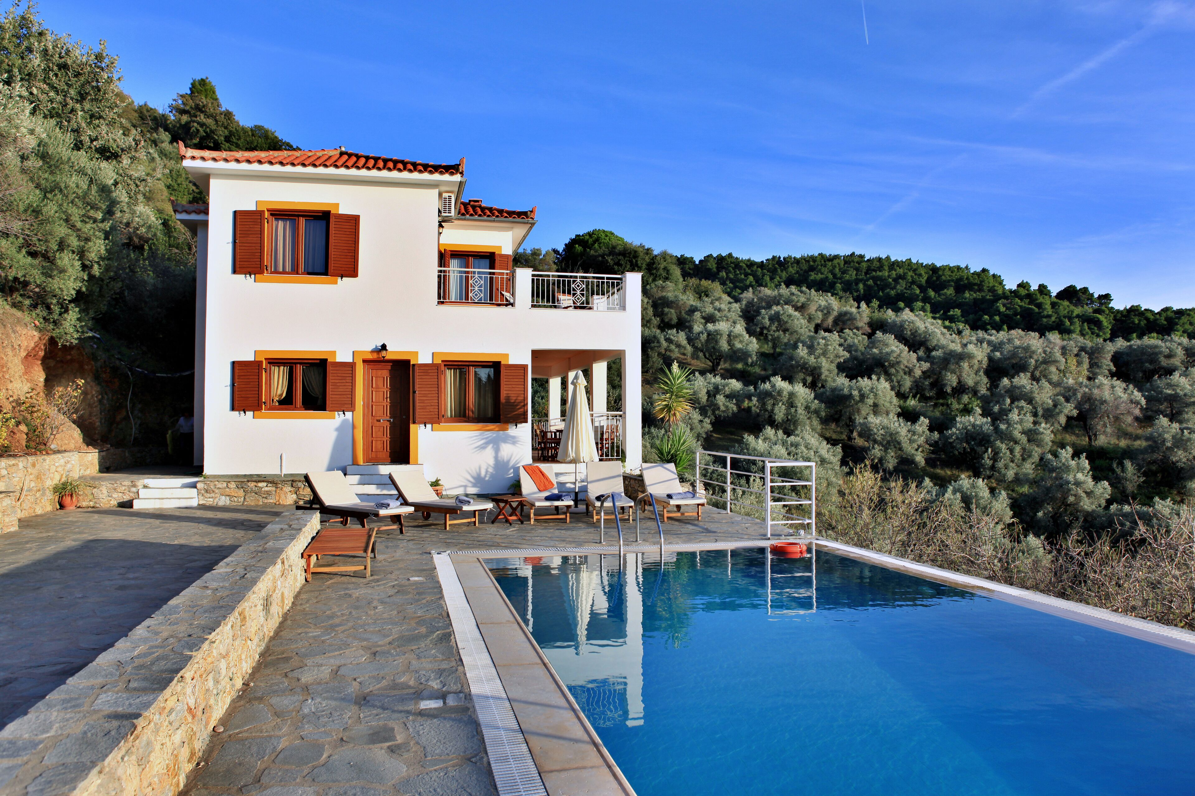 A two-story house with a swimming pool, outdoor seating, and a view of a forested hillside.