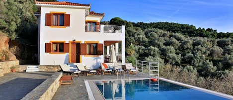 A two-story house with a swimming pool, outdoor seating, and a view of a forested hillside.