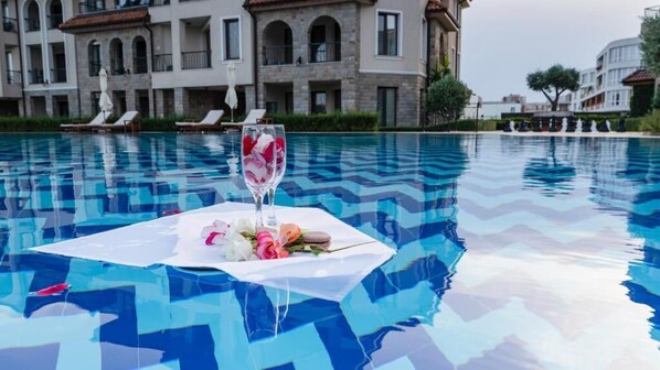 A pool with a glass of water and flowers on a tablecloth, surrounded by lounge chairs and a building in the background. - Burgas Beach Resort (Burgas)