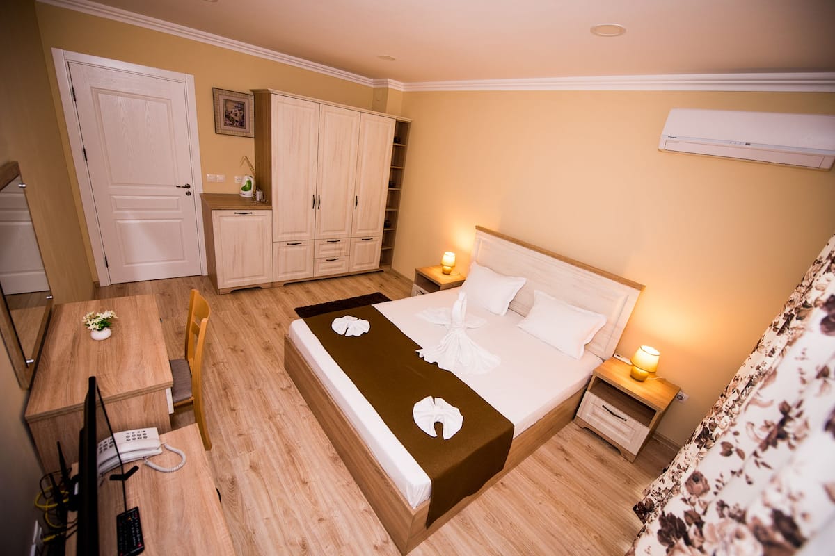 A hotel room with a bed, bedside tables, a desk with a telephone, a wardrobe, and an air conditioning unit.