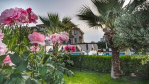 Um jardim com flores cor-de-rosa, uma palmeira e um prédio com piscina ao fundo.