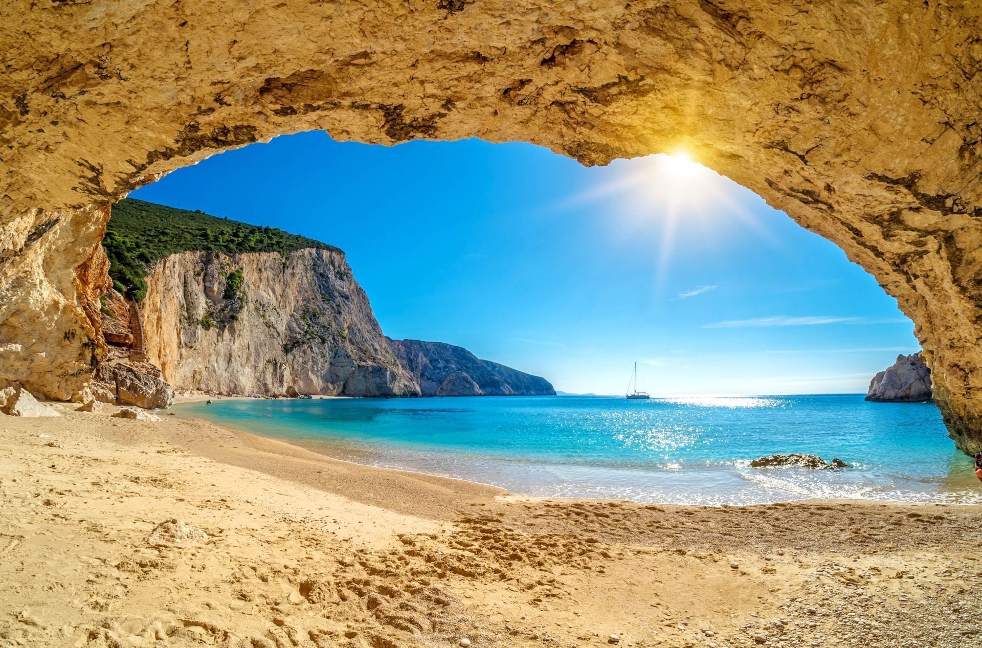 A beach with a natural rock arch formation, clear blue water, and a distant sailboat.