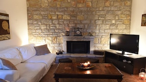 A living room with a stone fireplace, a white sofa, a wooden coffee table, and a flat-screen TV. - Village Villas Lefkada 2 floor villa, Pool, Large Garden near beaches, & city (Καριώτες)