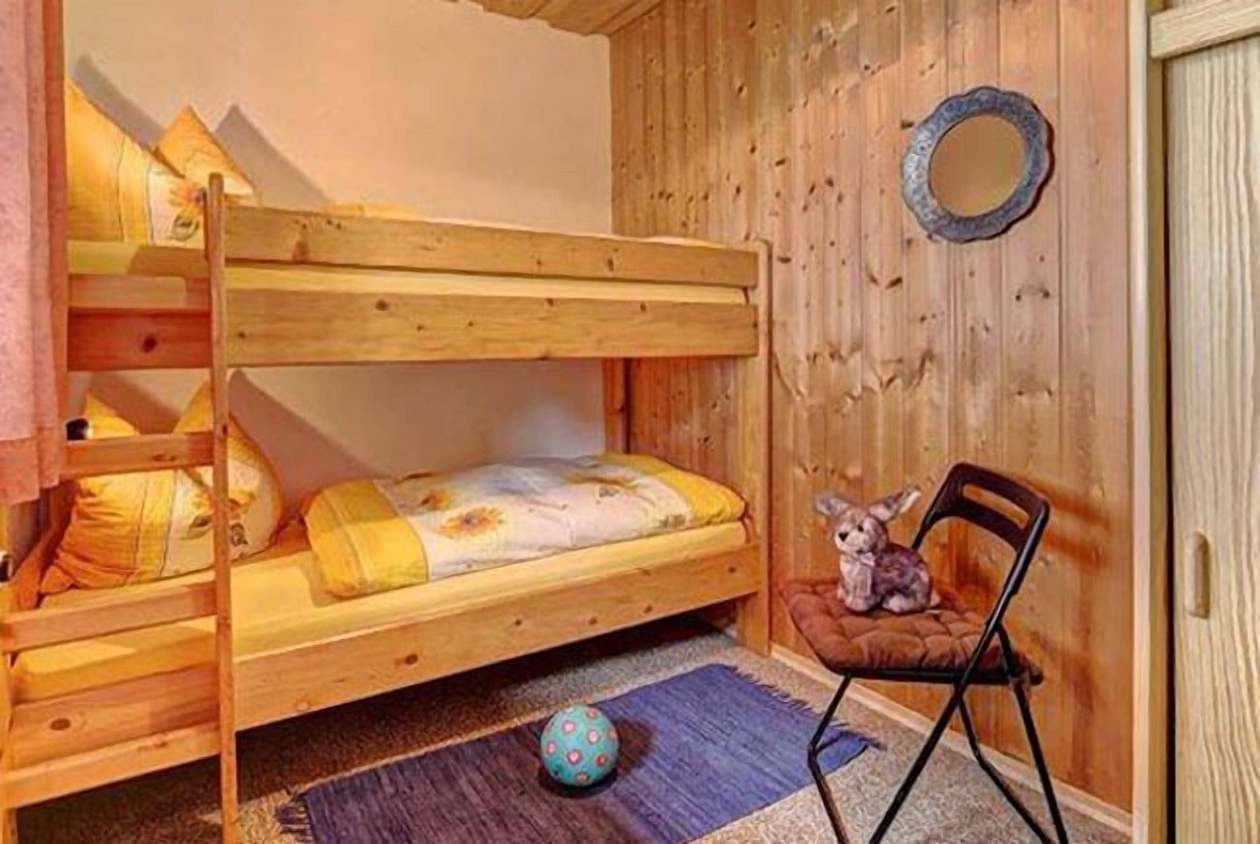 A bunk bed room with a wooden floor, a small chair, and a round mirror on the wall.