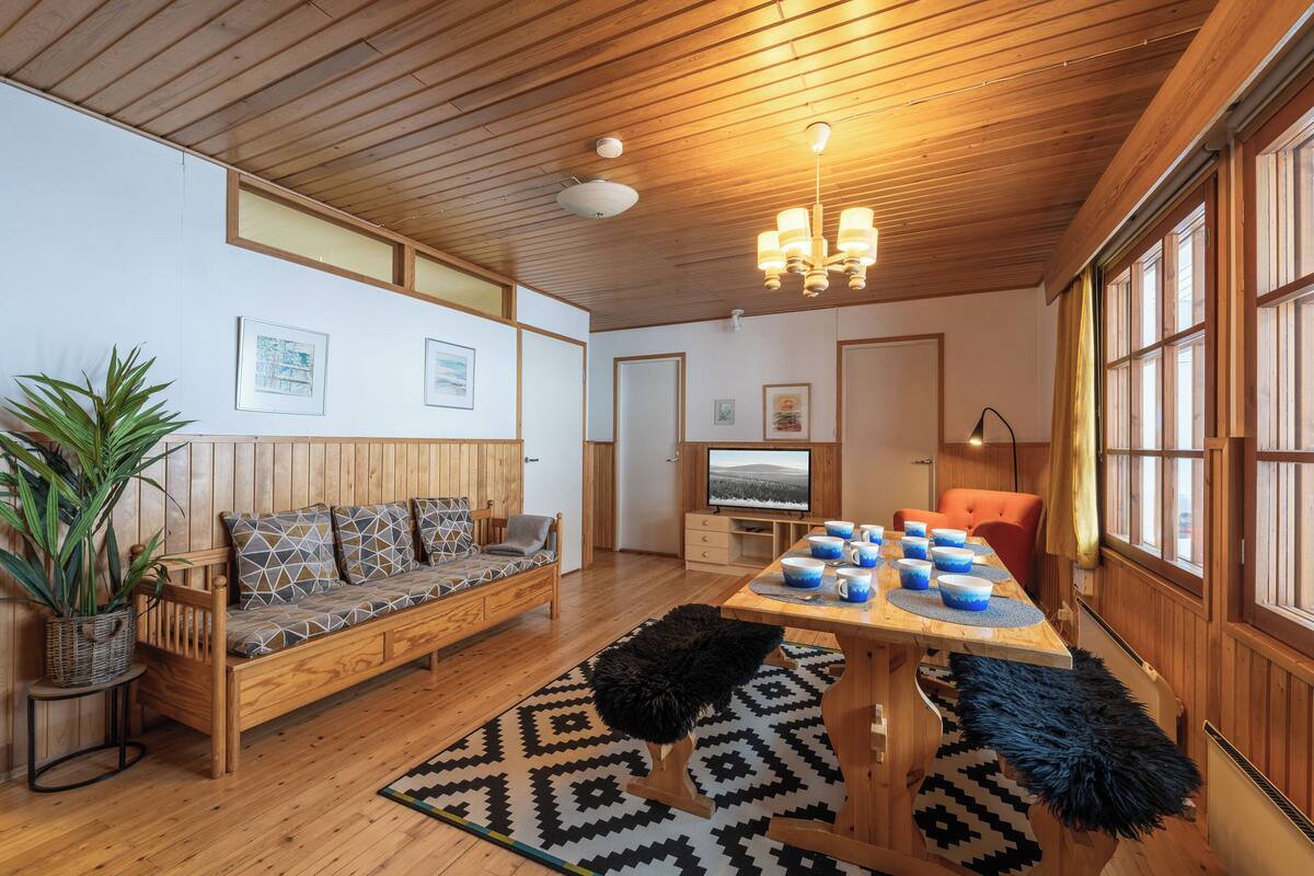 A cozy room with a wooden ceiling, a sofa, a dining table with blue cups, and a television.
