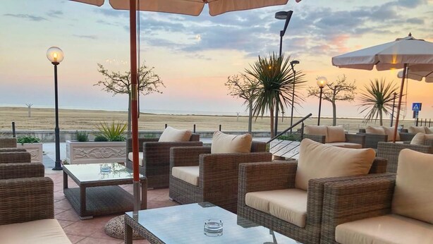 Een terras met rieten meubels en glazen tafels, met uitzicht op een strand bij zonsondergang.