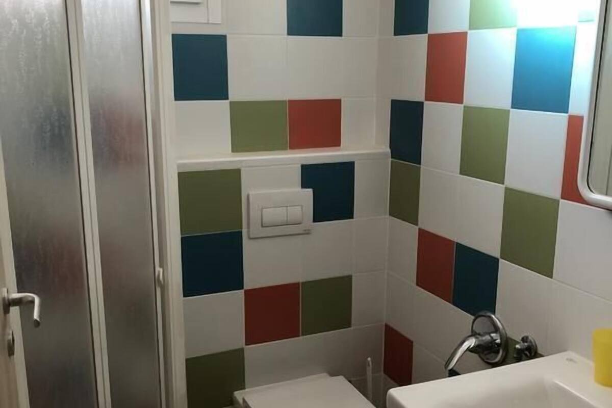 Une petite salle de bain avec des toilettes, un lavabo et un miroir. Les murs sont recouverts de carreaux disposés en motif à carreaux, avec des carrés verts, bleus et rouges.