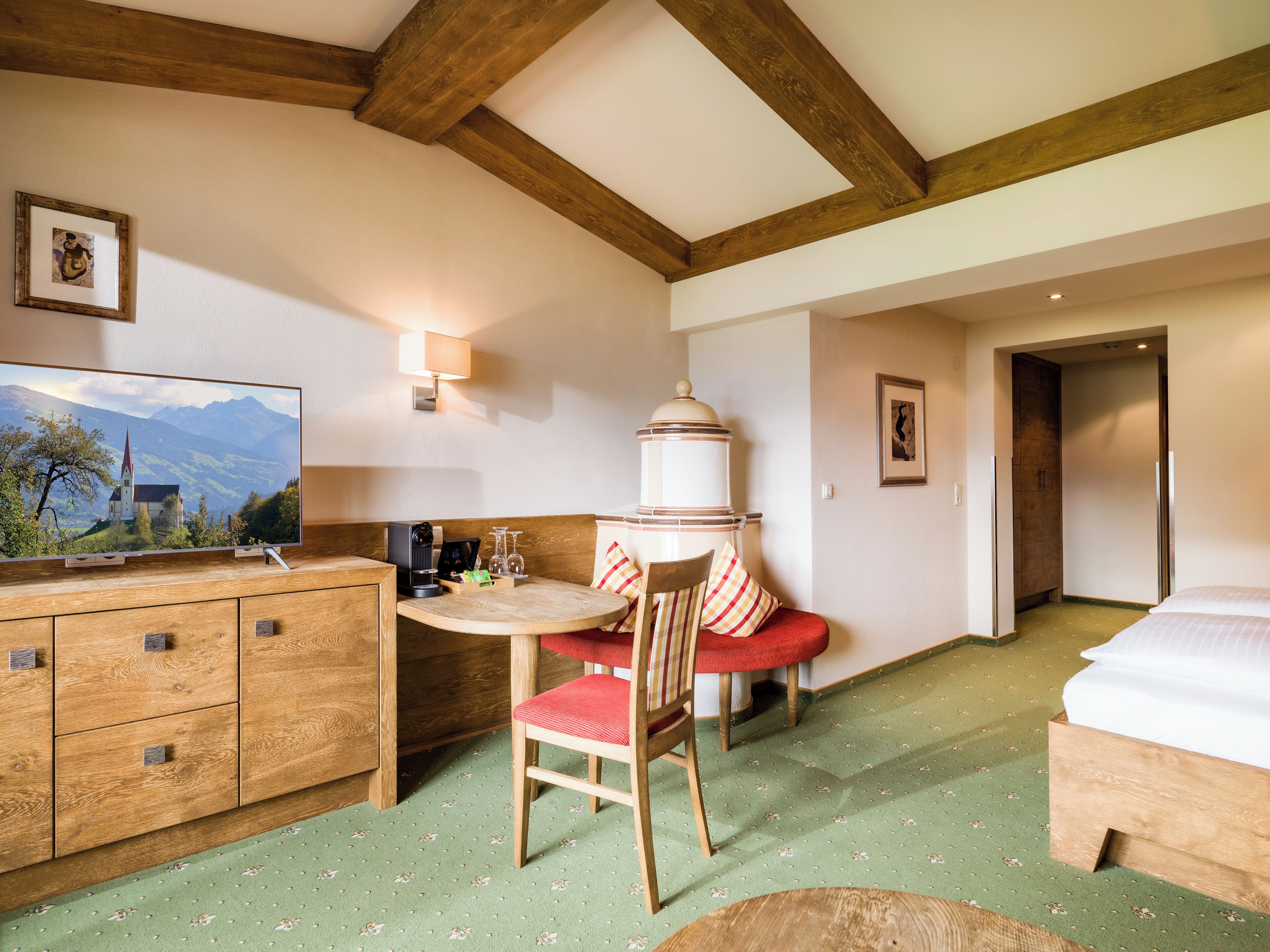 A hotel room with a wooden ceiling, a bed, a dining table with chairs, a small kitchen area, and a television displaying a scenic view.