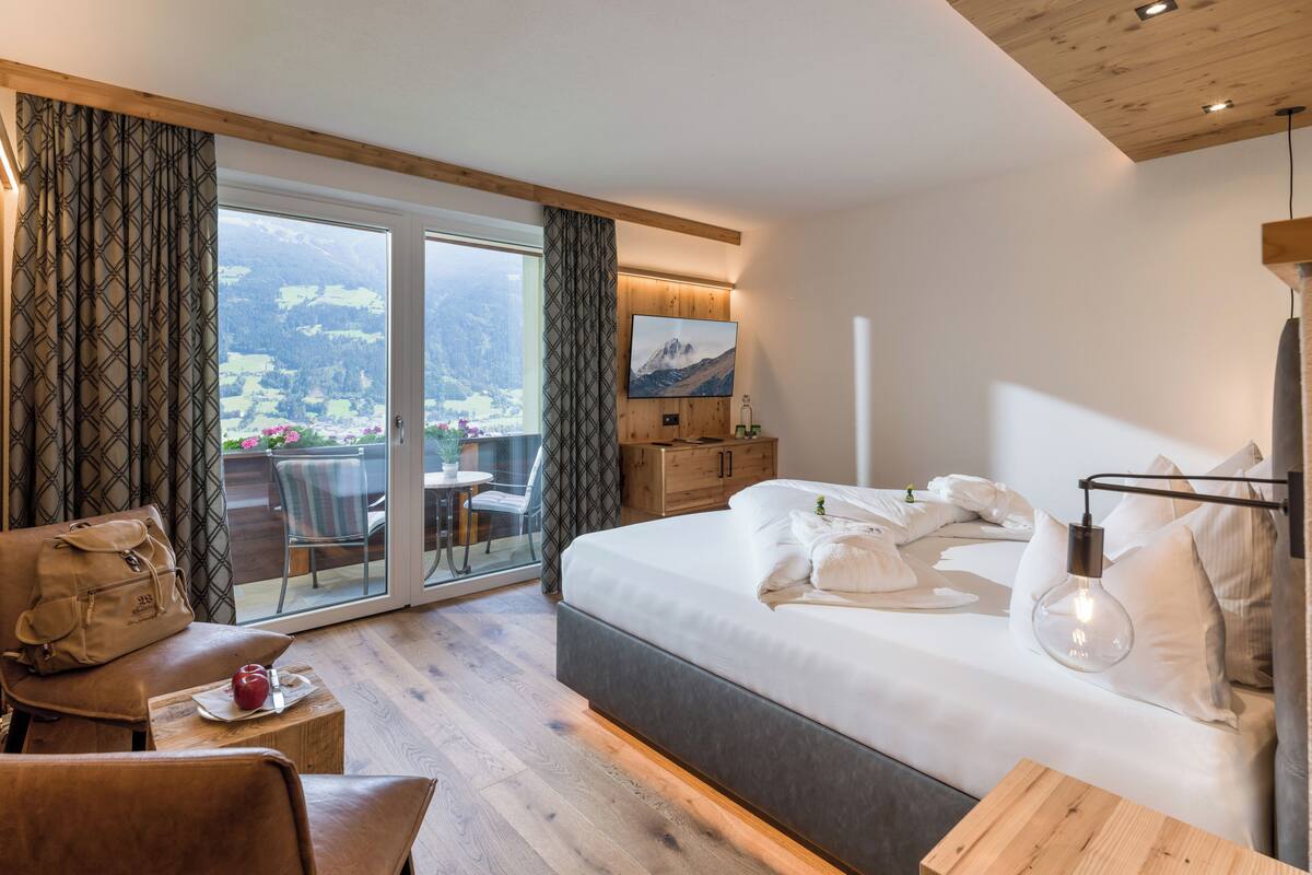 A hotel room with a bed, a desk, a chair, a handbag, and a view of a mountainous landscape through a sliding door.