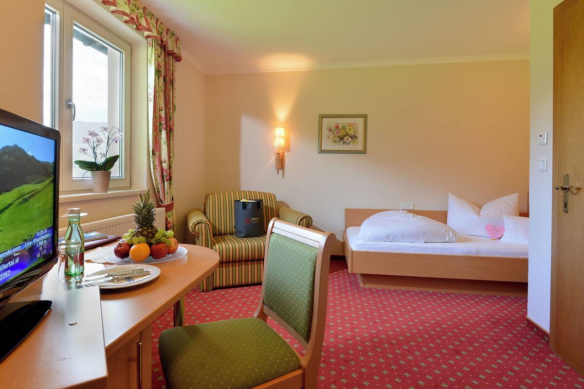 A hotel room with a bed, a desk with a computer, a chair, a small table with fruit, and a window with curtains.