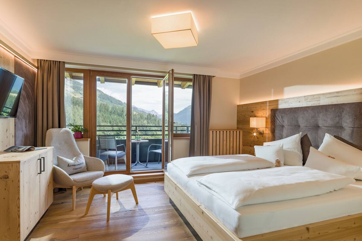 A hotel room with a wooden bed, a desk, a chair, a TV, and a balcony with a view of the mountains.