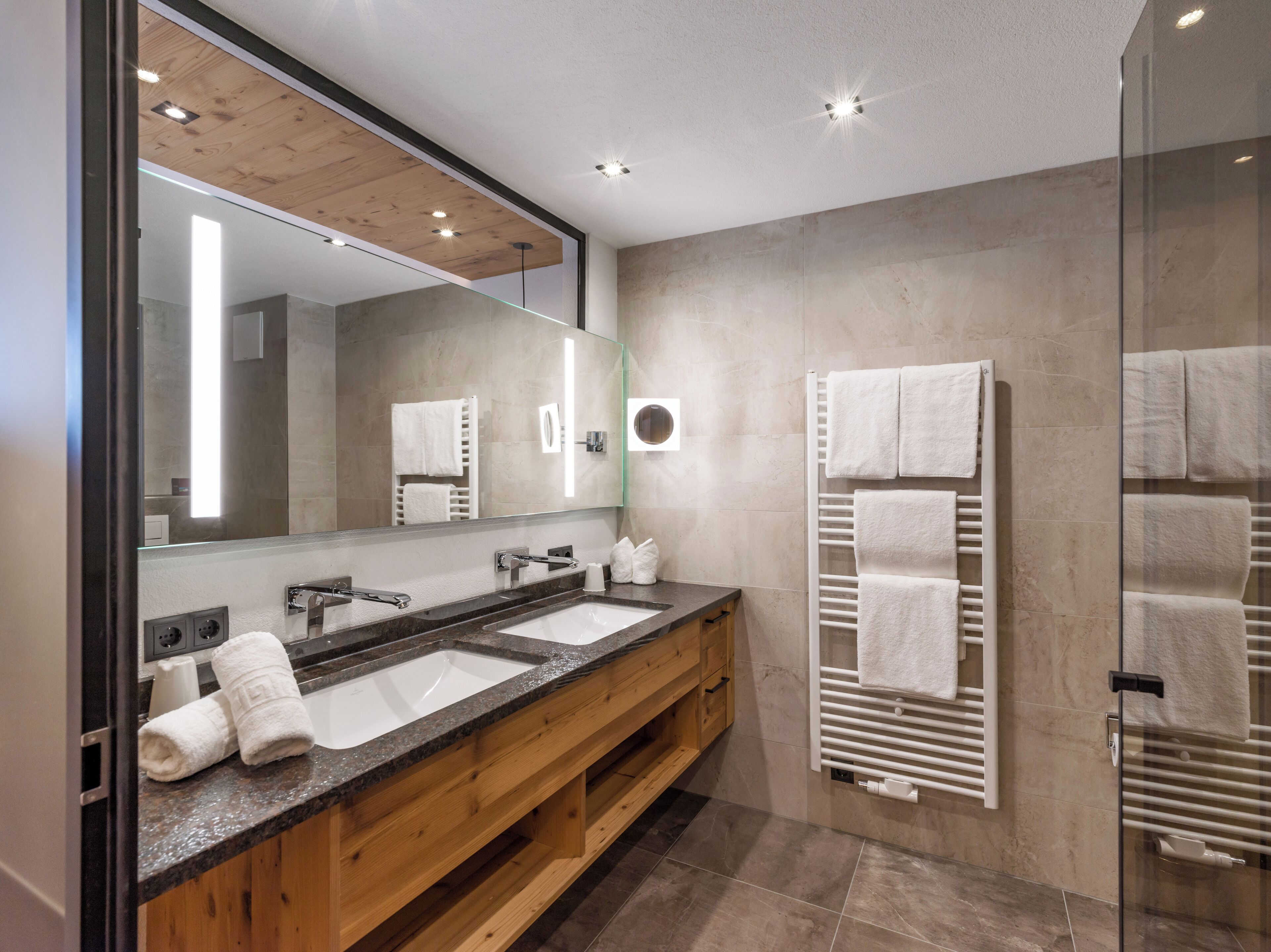 A modern bathroom with two sinks, wooden vanity, large mirror, towel rack, and recessed ceiling lights.
