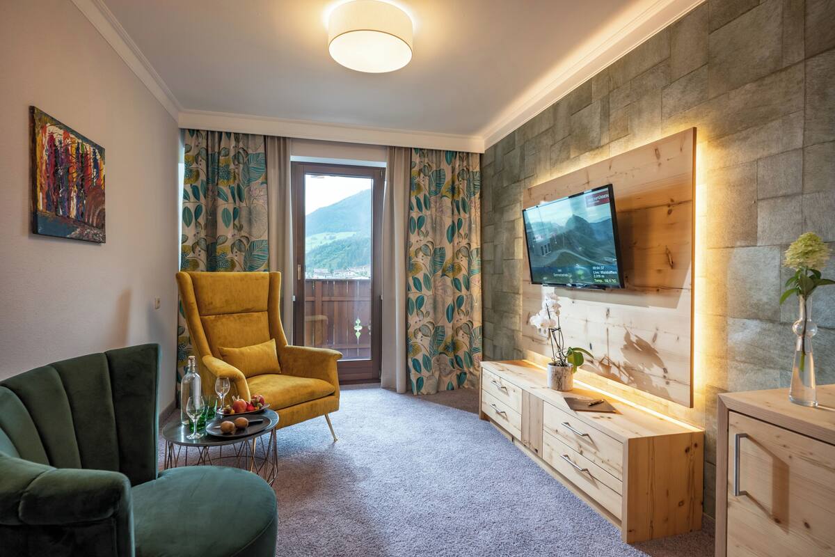 A modern hotel room with a large window, a yellow armchair, a green armchair, a wooden dresser, and a flat-screen TV mounted on the wall.