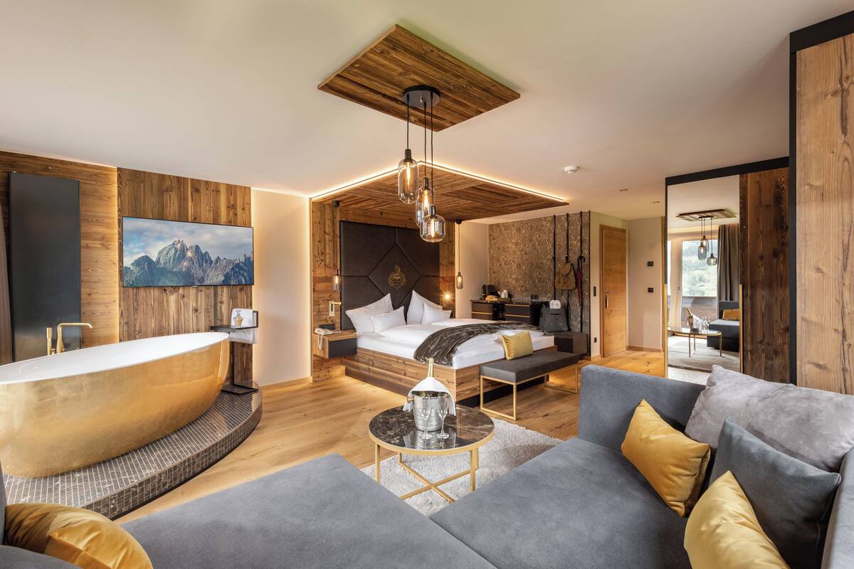 A modern hotel room with a large bathtub, a bed with a wooden headboard, a gray sofa, and a wooden ceiling.