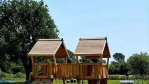 Una estructura de madera para juegos con dos zonas de escalada separadas y un tobogán, rodeada de un campo de césped y árboles.
