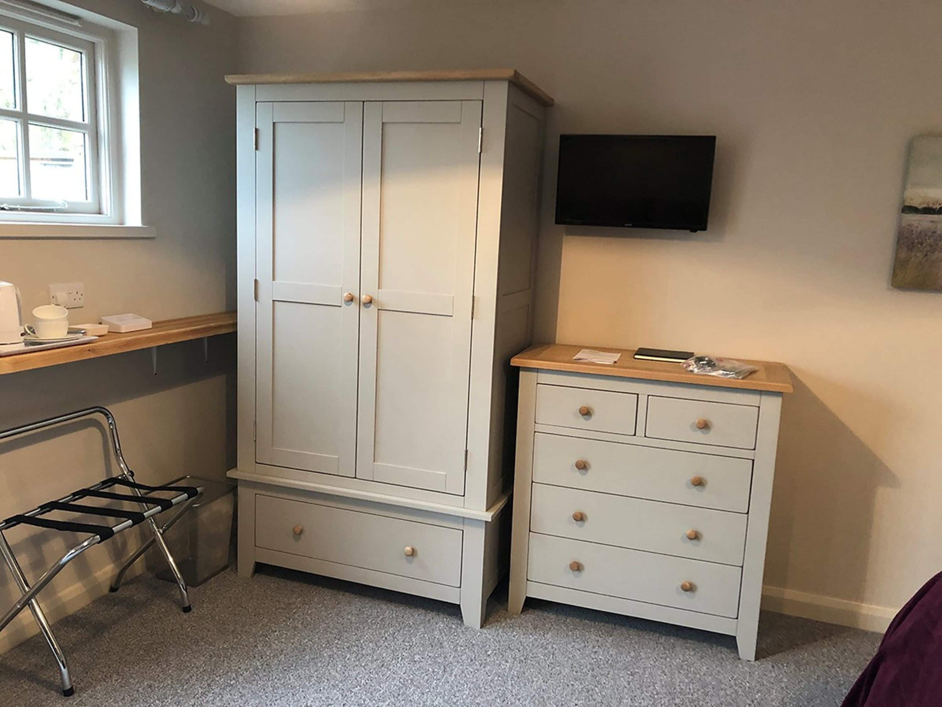 A room with a large white wardrobe, a small white dresser, a metal drying rack, and a flat-screen TV mounted on the wall.