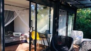 A room with a bed, a desk, and a chair. There is a glass door leading to an outdoor area with wicker furniture. - Area 4 eco cubes (Ella)