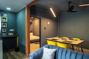 A modern room with a blue sofa, a wooden desk, a dining table with yellow chairs, a ceiling fan, and a wall-mounted light fixture. - Area 4 eco cubes (Ella)