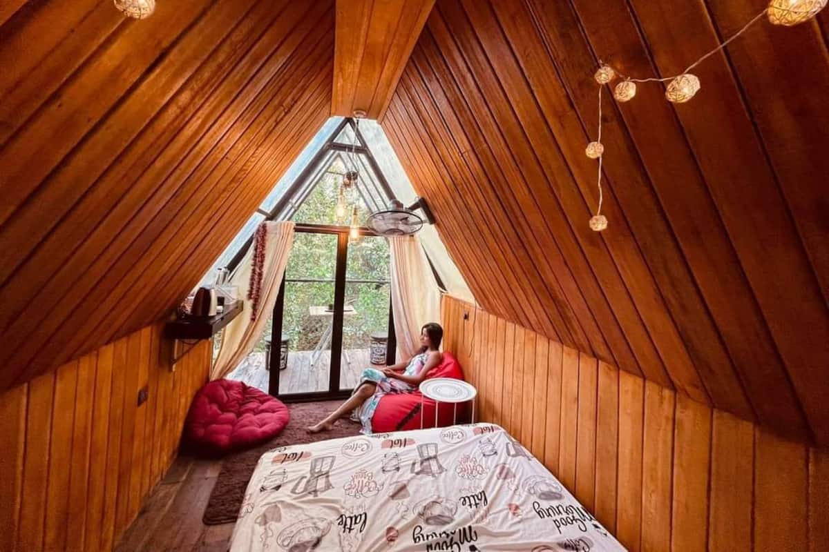 A cozy attic bedroom with wooden walls, a sloped ceiling, and a large window. There is a bed with a patterned bedspread, a red bean bag chair, and a small round table.