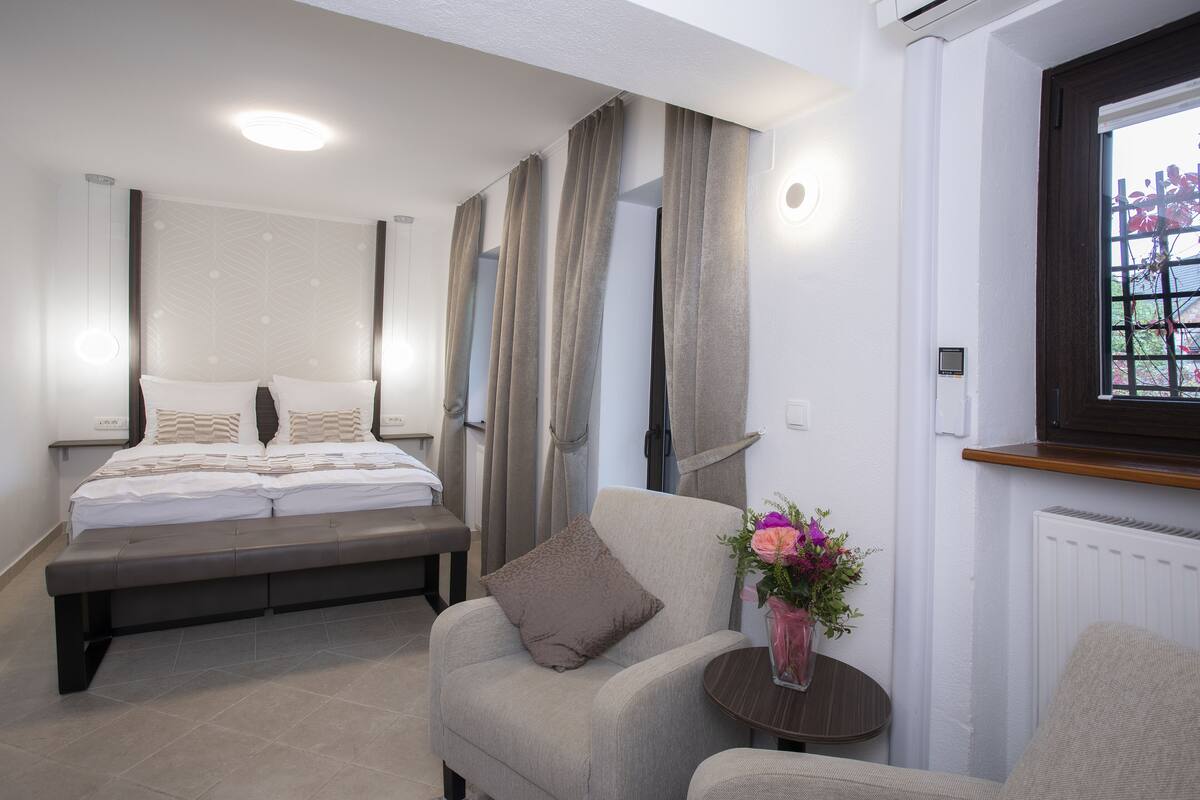 A bedroom with a bed, a chair, a small table with a vase of flowers, and a window with a view of trees.