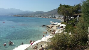 Vista costera con aguas cristalinas, una playa con sombrillas y un pueblo al fondo.