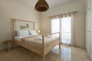 A bedroom with a wooden bed, a small stool, a lamp, and a large window with curtains.