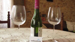 Un tavolo con una bottiglia di vino e due bicchieri, una tovaglia di pizzo e un muro in pietra con quadri appesi.