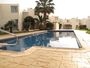 A swimming pool with a tiled deck, surrounded by palm trees and modern buildings. - Webooking Pegia Apartment (Pegeia)