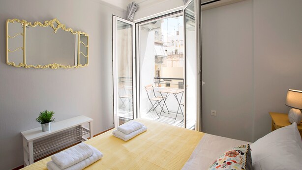 A hotel room with a bed, a bedside table with a lamp, a mirror, and a sliding glass door leading to a balcony with a table and chairs. - Attractive Flat near the Acropolis Museum - Athenian Homes, Athenian Homes (Athens)